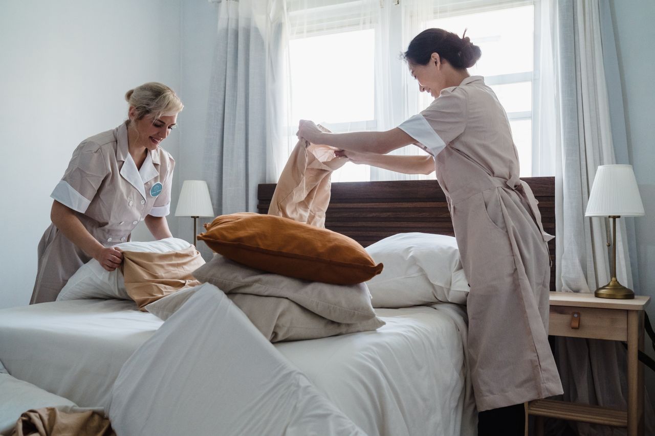 journey Two housekeepers in uniform fixing a hotel room bed with fresh linen and pillows.